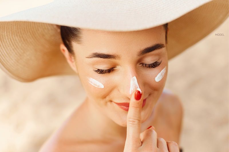 Woman applying sunscreen to her nose under a hat.