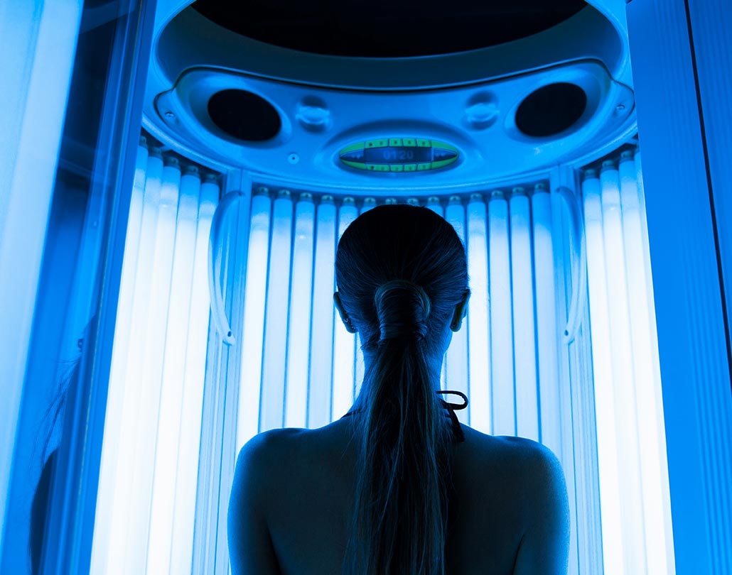 A dark silhouette of a woman stands in front of the ultraviolet blue glow of a tanning bed.
