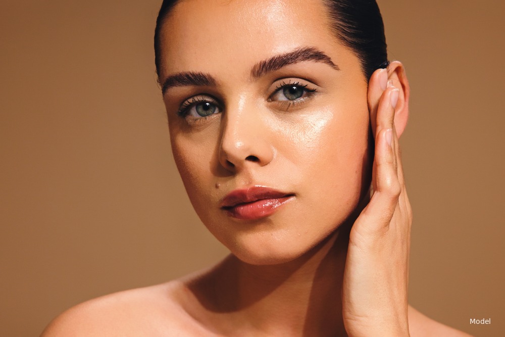 Woman with dewy glowing skin, hand near face, hair pulled back, warm studio background